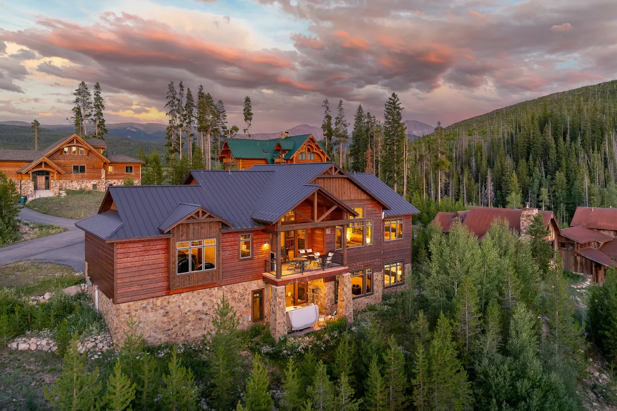 This exterior image displays the main house with expansive windows set against a backdrop of trees, mountains, and a colorful sunset sky. The inviting balcony hints at relaxation, while the surrounding landscape suggests opportunities for nature walks and cozy evenings by a fire pit.