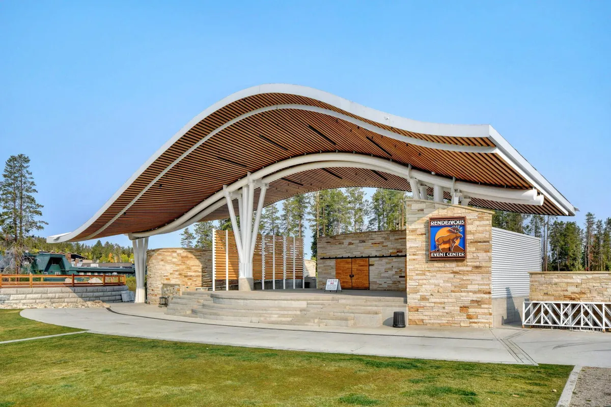 An exterior image featuring a unique curved-roof structure and stone facade, with an open stage area ready for concerts and community events. The scene suggests a festive, communal atmosphere perfect for gatherings, music, and cultural celebrations under the open sky.