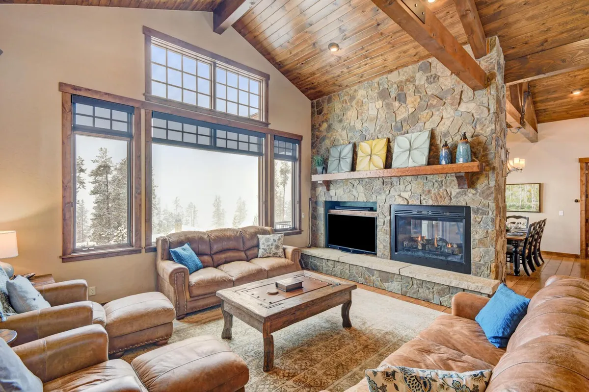 A spacious, welcoming living area with a grand stone fireplace, large windows framing scenic views, wooden beams, and comfortable seating. The setting invites relaxation and socializing, perfect for enjoying gourmet coffee or unwinding after a day of adventure.