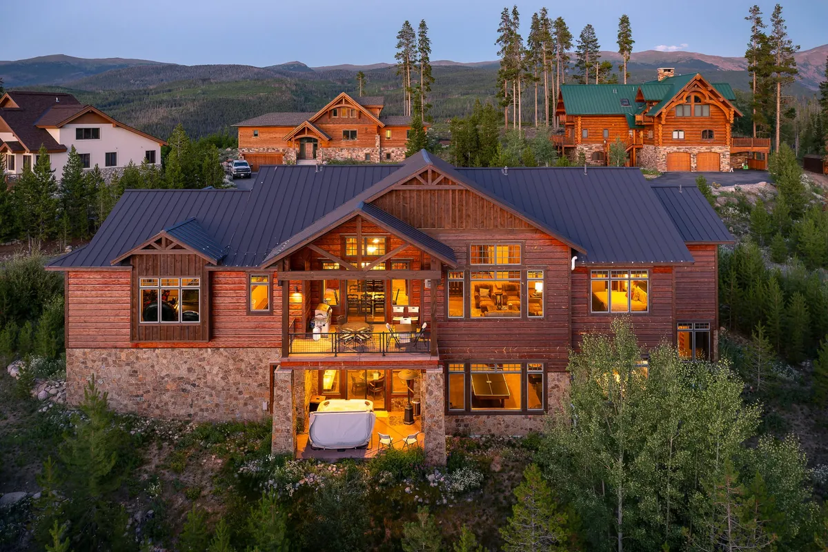 The hero image showcases a stunning timber home nestled among tall pine trees, with expansive windows reflecting the sunset. The centerpiece is a steaming outdoor hot tub set on a spacious patio, inviting guests to relax while taking in panoramic mountain views. Cozy seating surrounds a crackling fire pit, enhancing the atmosphere of comfort and luxury amid a serene alpine setting.