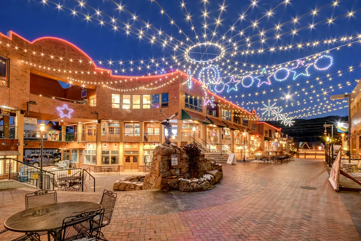A lively exterior scene of downtown at night: string lights twinkle above festive outdoor seating, decorative stars hang, and a stone fountain is the centerpiece. People can be seen dining, shopping, and socializing, giving the scene a joyful, community vibe.