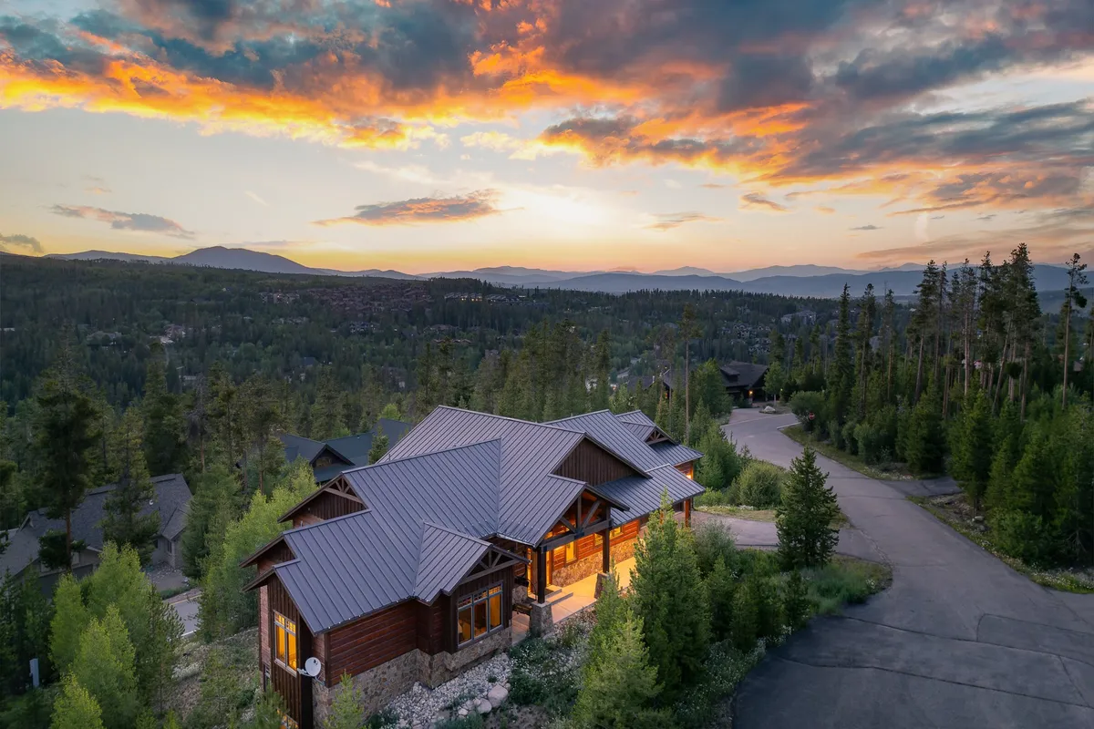 An inviting exterior view of the property showcases wooden architecture surrounded by trees, with a tranquil sunset sky and a driveway leading up. The spacious deck hints at areas for outdoor play, relaxation, and family gatherings, set against the natural landscape.
