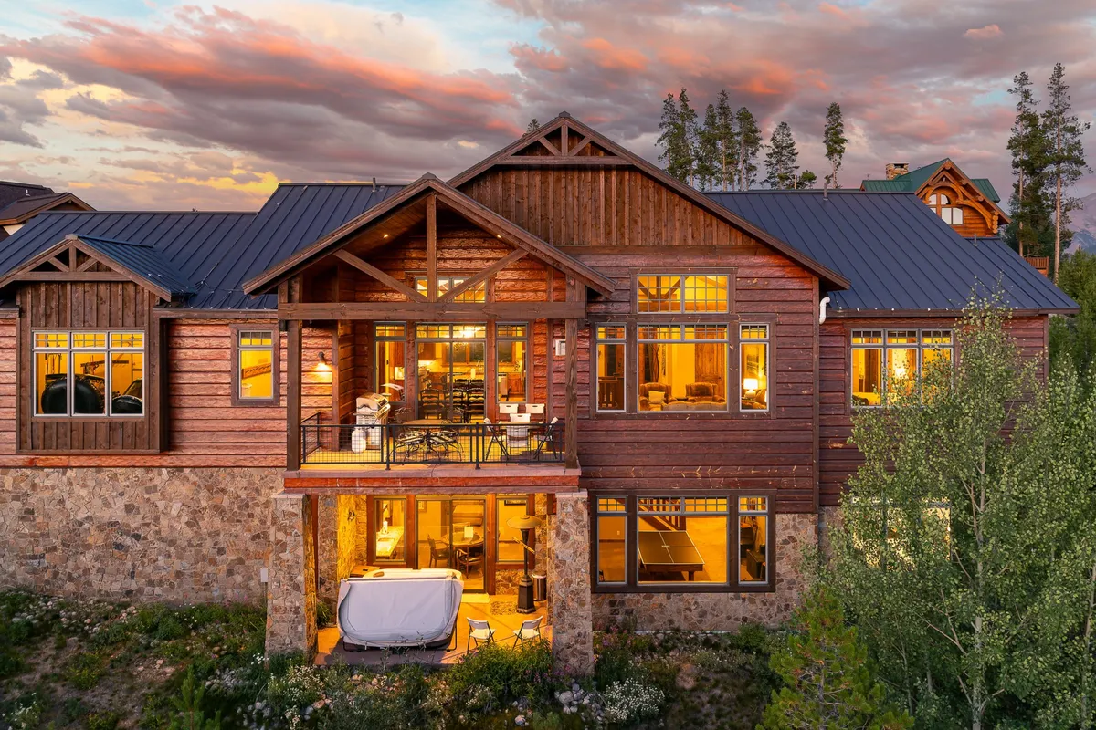 This exterior image shows a beautifully designed wooden home with large windows, an inviting outdoor seating area, and a landscaped garden. The stone foundation and cozy patio are perfect for gatherings, while the sunset sky adds a touch of magic to the setting.