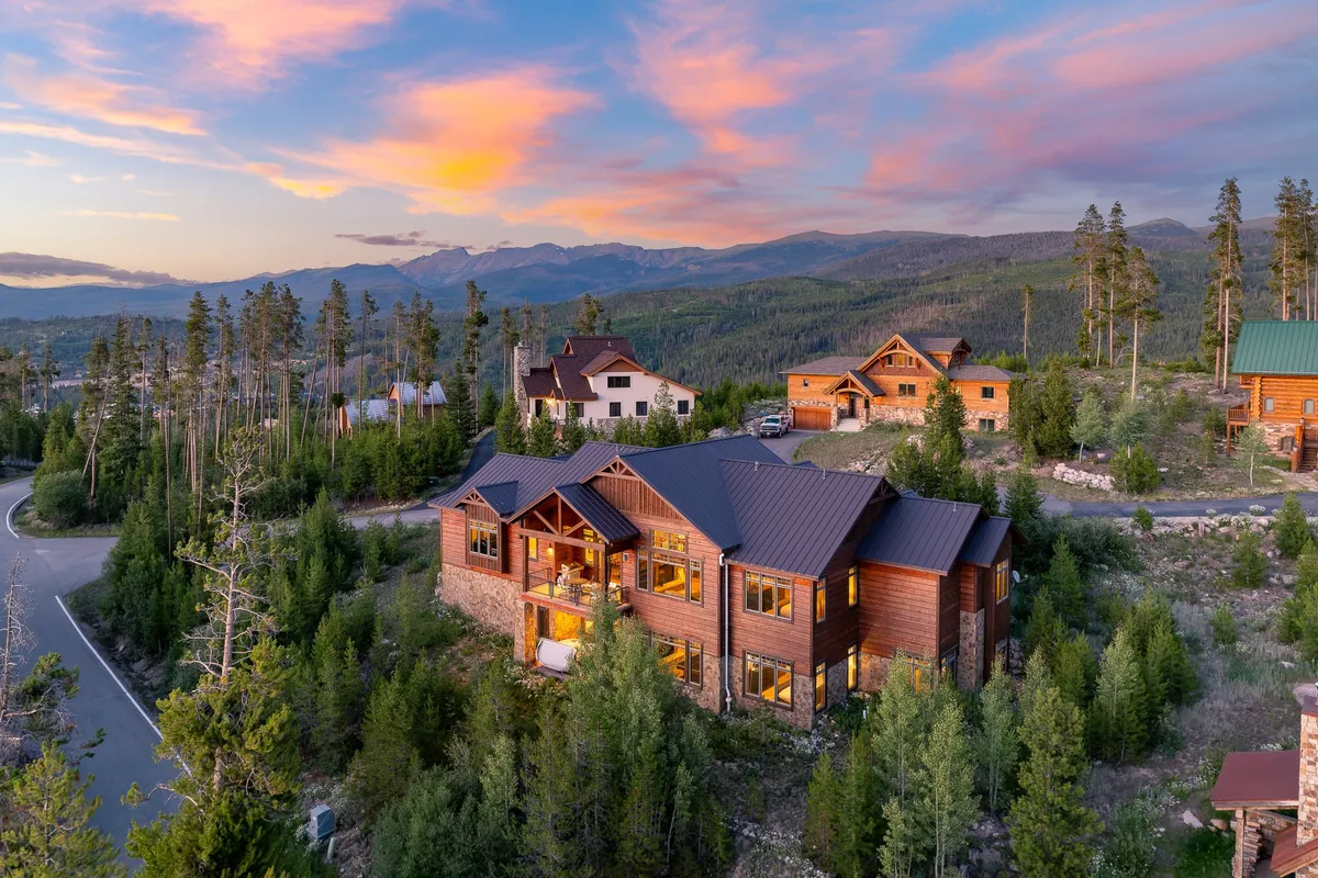 This image shows the main house nestled among trees, with a backdrop of stunning mountains and a vivid sunset sky. The scene conveys a sense of serenity, natural beauty, and proximity to outdoor adventure. The house appears inviting, with a spacious porch that hints at relaxation and gatherings after a fun day out.