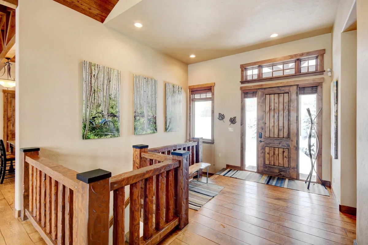 A bright, light-filled entrance area featuring a wooden front door, light-colored walls, nature-themed artwork, and a wooden railing. The space is airy and inviting, projecting a clean and tranquil environment.