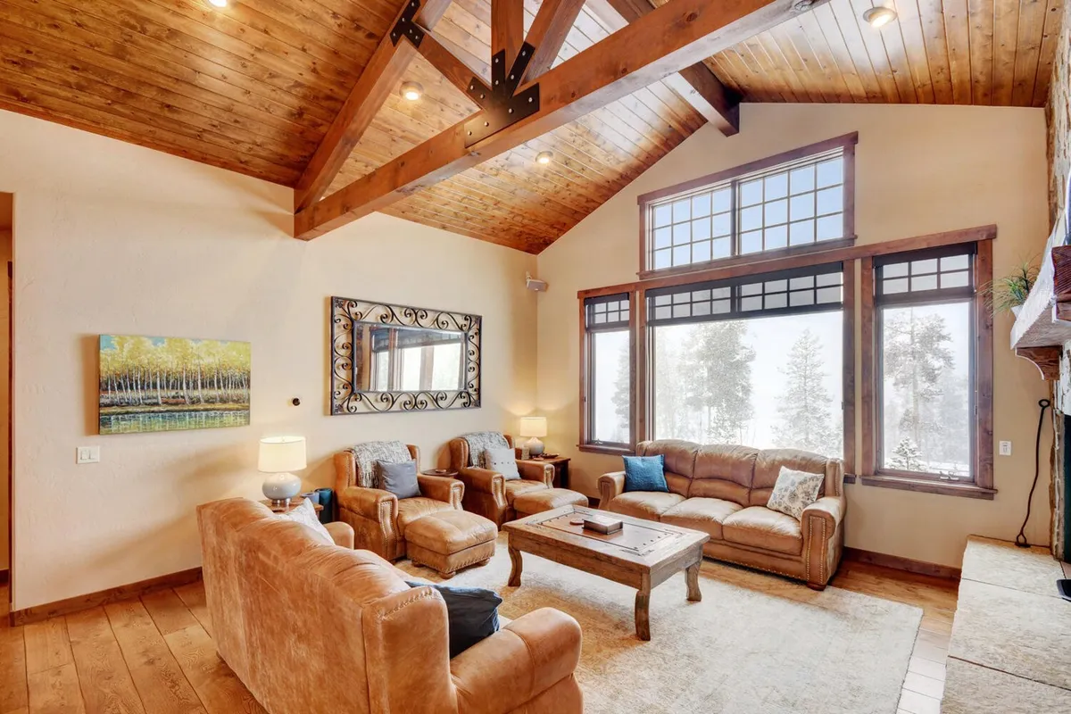 A warm, welcoming living room interior with rustic wooden beams, large windows framing outdoor views, a comfortable seating area, and a cozy fireplace. Snow gently falls outside, visible through the windows, enhancing feelings of relaxation and connection to nature.