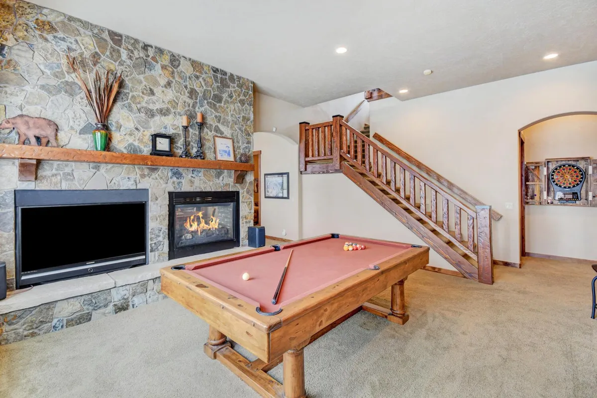 A spacious rec room featuring a pool table at the center, a stone fireplace glowing warmly in the background, a rustic stone wall, and a staircase leading upward. The room is arranged for both games and cozy relaxation, with seating areas ideal for socializing or movie nights.