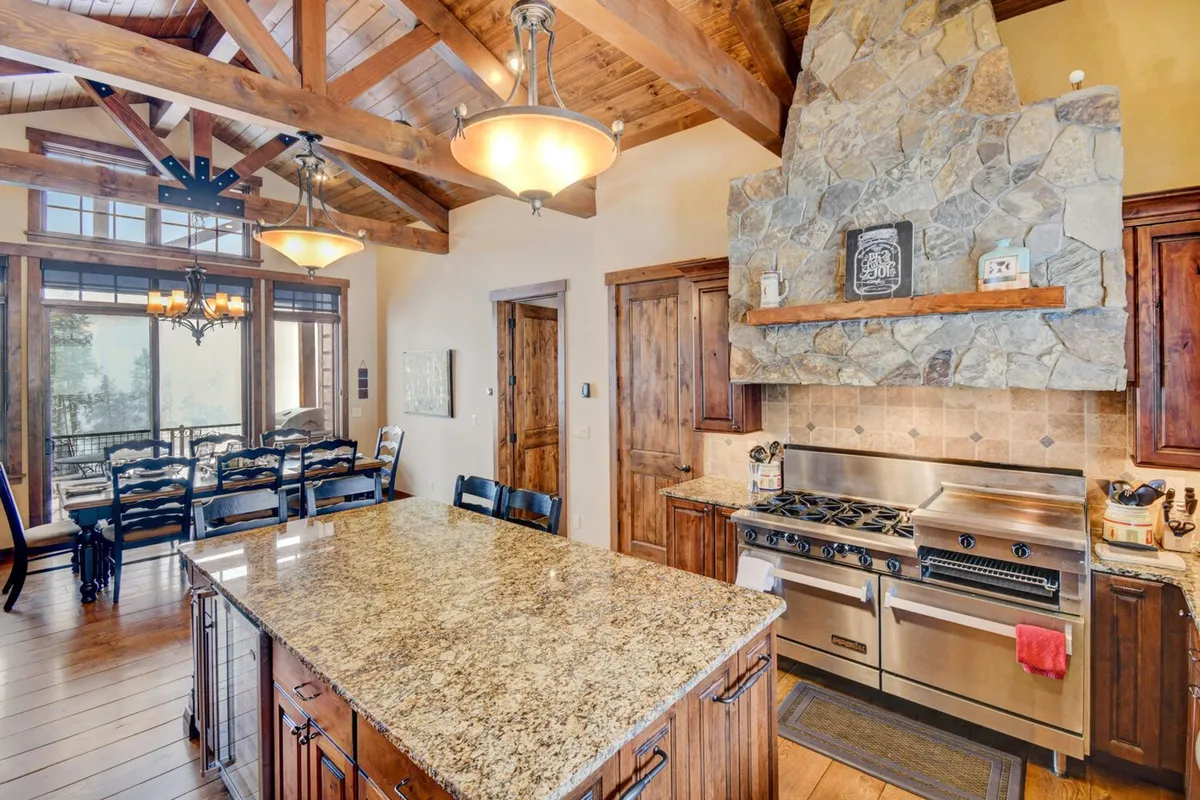A chef’s kitchen and dining area highlighted by a large granite island, high-end stainless appliances, and wooden beams overhead. The stone fireplace in the background ties the kitchen to the living space, while the generous dining table invites shared meals and celebrations. Designer touches and ample space showcase both culinary and social possibilities.