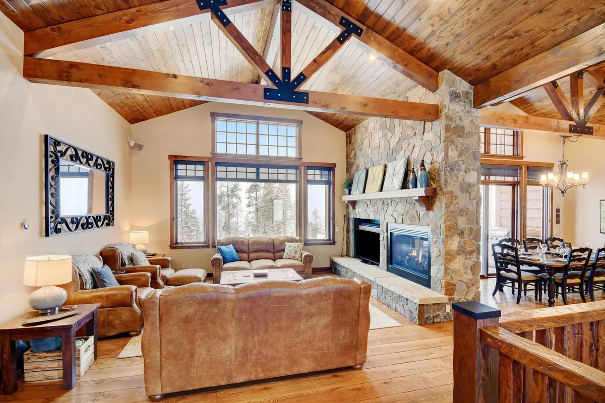A grand, open-plan living space featuring soaring vaulted wooden ceilings, a striking stone fireplace, and wall-to-wall panoramic windows that frame breathtaking mountain views. The comfortable seating area, accented by plush designer furnishings, flows seamlessly into a dining area with a large table set for a gathering. Warm, radiant lighting and wood textures create an inviting atmosphere ideal for both relaxation and celebration.