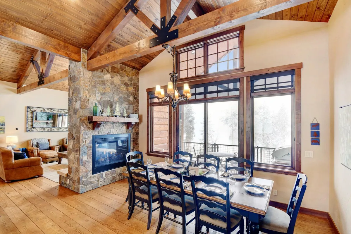 A cozy dining space centered around a large table placed by expansive windows and a glowing fireplace. Wooden beams and tastefully selected furnishings set the stage for warm family gatherings, communal celebrations, and enjoying the view of the snowy outdoors. The communal mood is palpable, inviting guests to linger and connect.