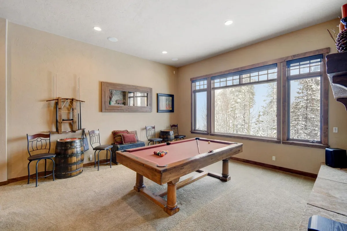 A bright and casual lower-level game room with a pool table, large windows looking out onto scenic views, a wooden barrel accent, and a comfortable seating area. Sunlight streams in, creating a friendly, social environment perfect for all ages.