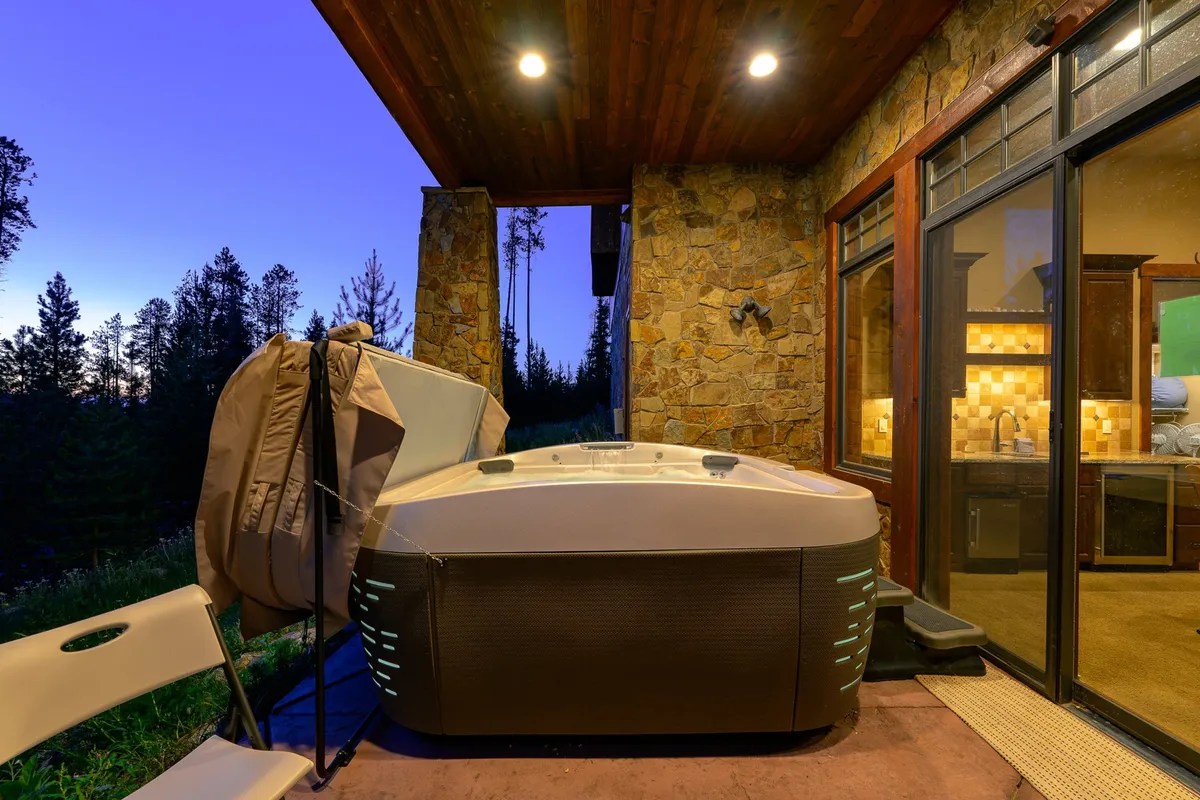 An inviting exterior patio with a steaming hot tub set beneath a wooden ceiling and stone wall, surrounded by tranquil trees. The scene is perfect for relaxing after a day of adventure—guests can soak under the stars while enjoying the peaceful natural setting.